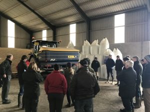 The Soil & Nutrient Network group standing inside a shed discussing soil improvers and artificial fertilisers. They are standing next to a tractor and fertiliser spreader