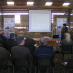 A presentation being given in a farm shed. A group of people seated, watching and listening to a person leading a discussion with a whiteboard behind them showing a power point presentation.