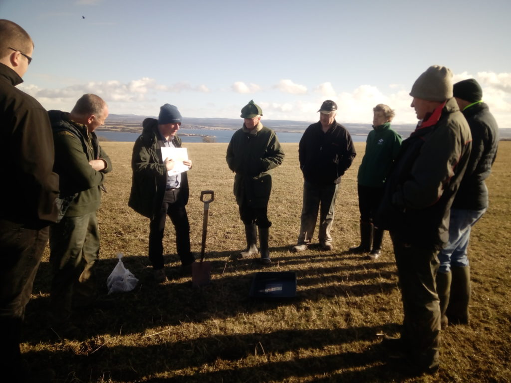 A group of farmers at the Inverness-shire Soil & Nutrient Network meeting, standing in a circular formation in a field with a low sun casting long shadows