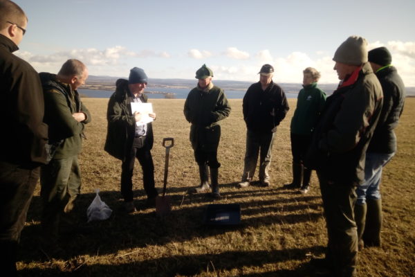 A group of farmers at the Inverness-shire Soil & Nutrient Network meeting, standing in a circular formation in a field with a low sun casting long shadows