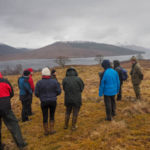 A group of people standing in an upland area near Lochaber with a cloudy sky and mountains in the backgroud over a loch