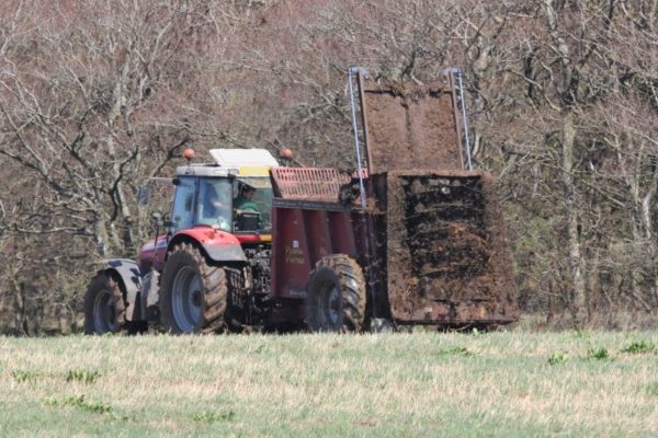 muck spreader in action in grassland field with trees in the background