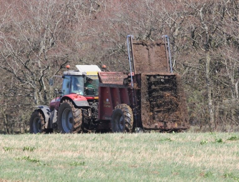 muck spreader in action in grassland field with trees in the background