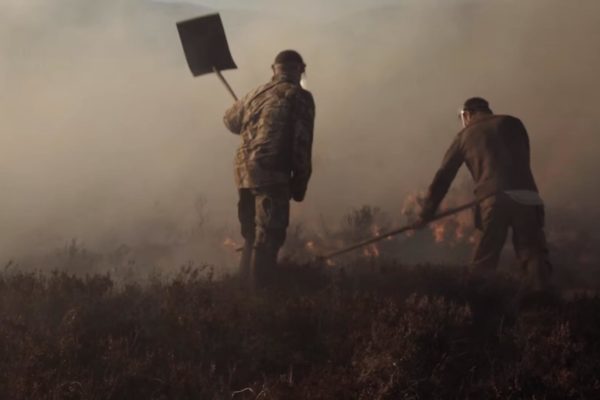 Photo showing two men beating burning moorland with fire paddles. The fire is not visible but in front of the men is dense smoke.