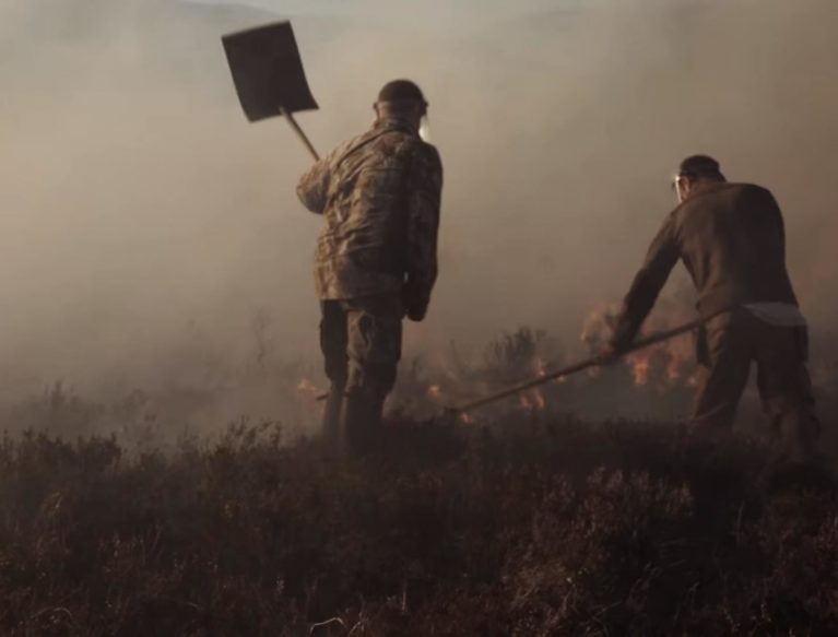 Photo showing two men beating burning moorland with fire paddles. The fire is not visible but in front of the men is dense smoke.