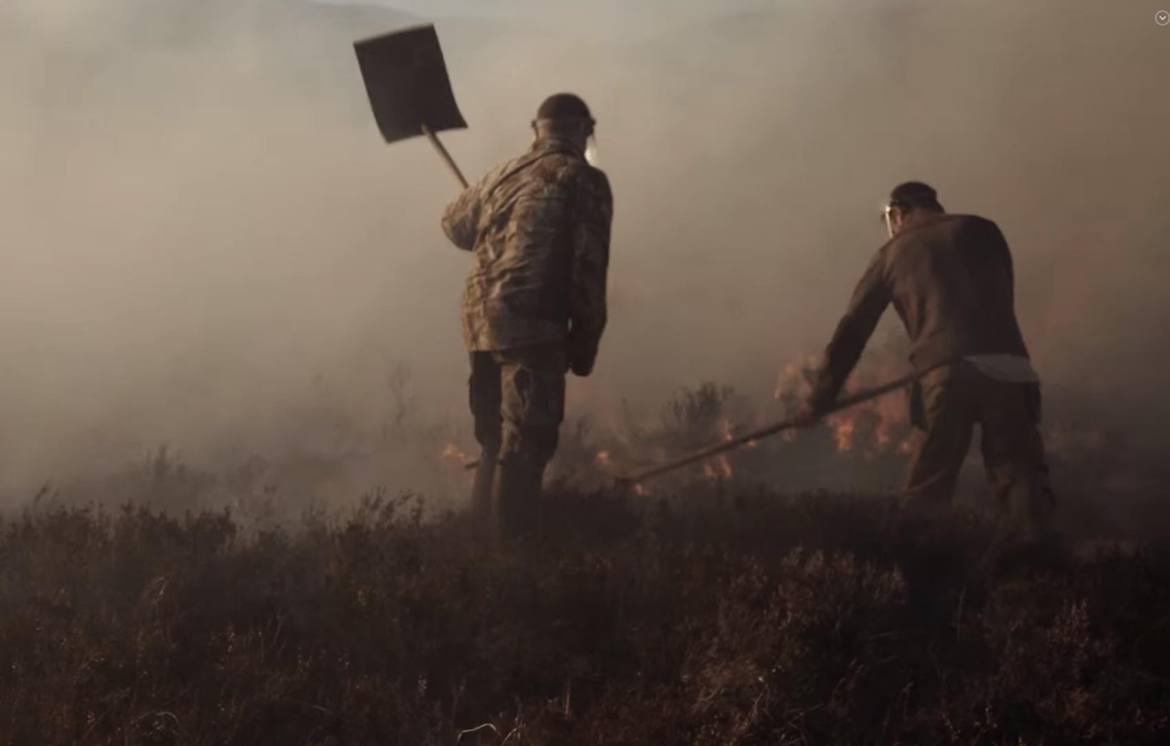 Photo showing two men beating burning moorland with fire paddles. The fire is not visible but in front of the men is dense smoke.