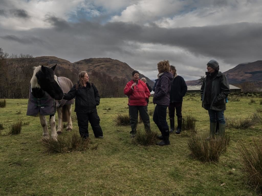 Five women and a pony stand in a grassy field with mountains in the background