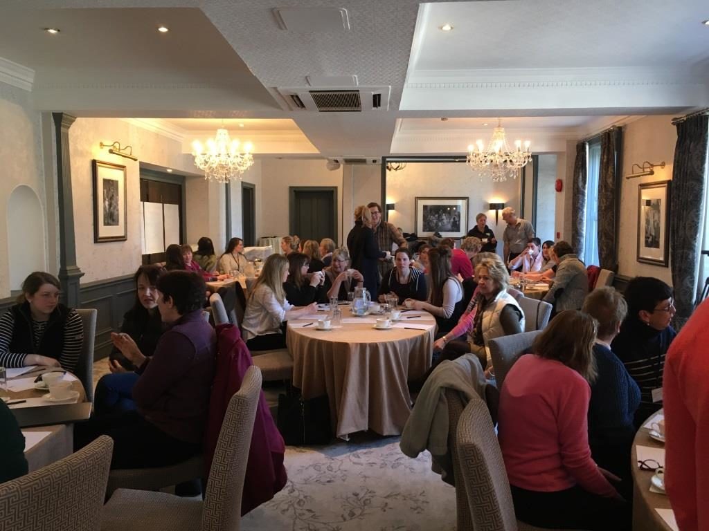 Many women sit around tables in a conference room, discussing animatedly