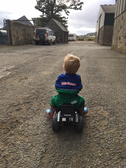 Small child riding a toy tractor wearing a boilersuit with the words ' Little Farmer' on it