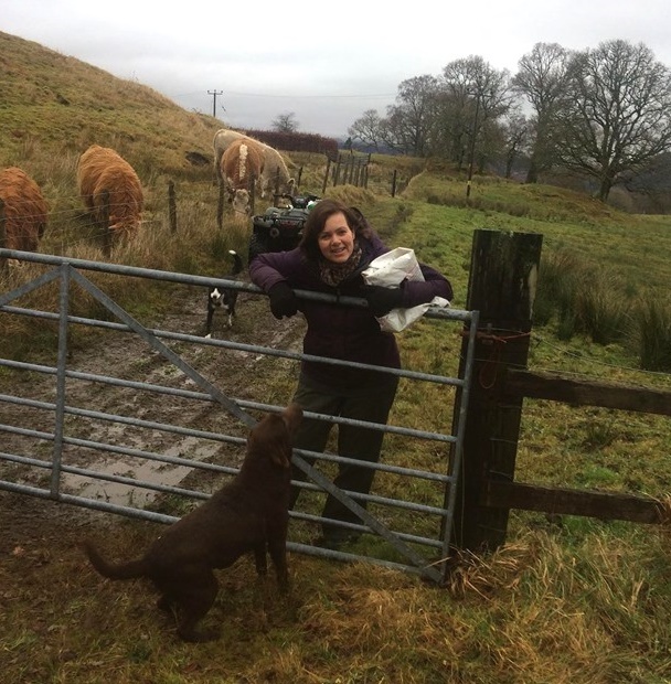 Woman holding a bag of feed, leaning over a gate with dogs at her feet and cattle in the background