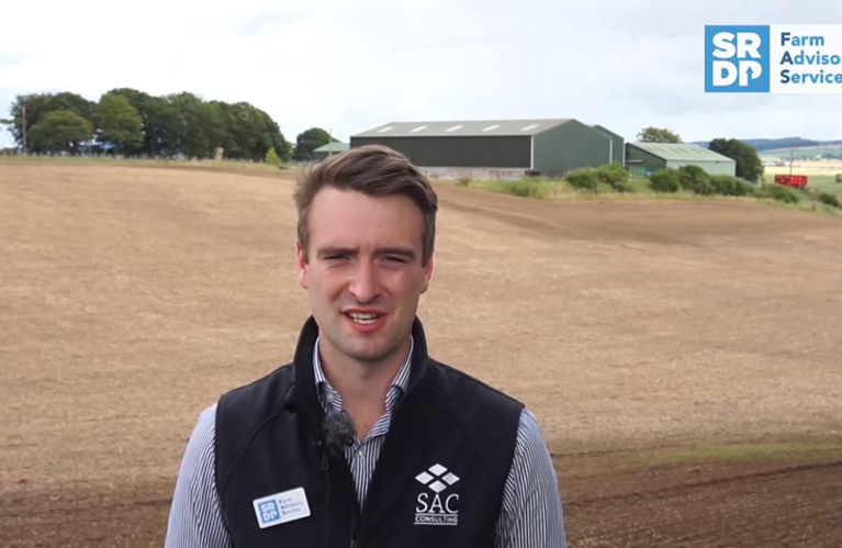 A young man looking towards the camera in his work uniform/gillet. He's standing in a recently worked arable field that has some recently planted crop starting to germinate. There is a farm steading in the background.
