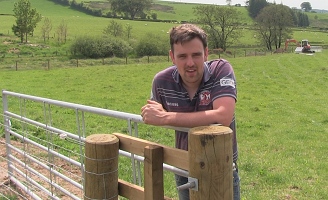 Young man looking towards the camera whilst leaning on a gate and gate post in a green field. There is a fence in the background with some trees and a digger in the distance.