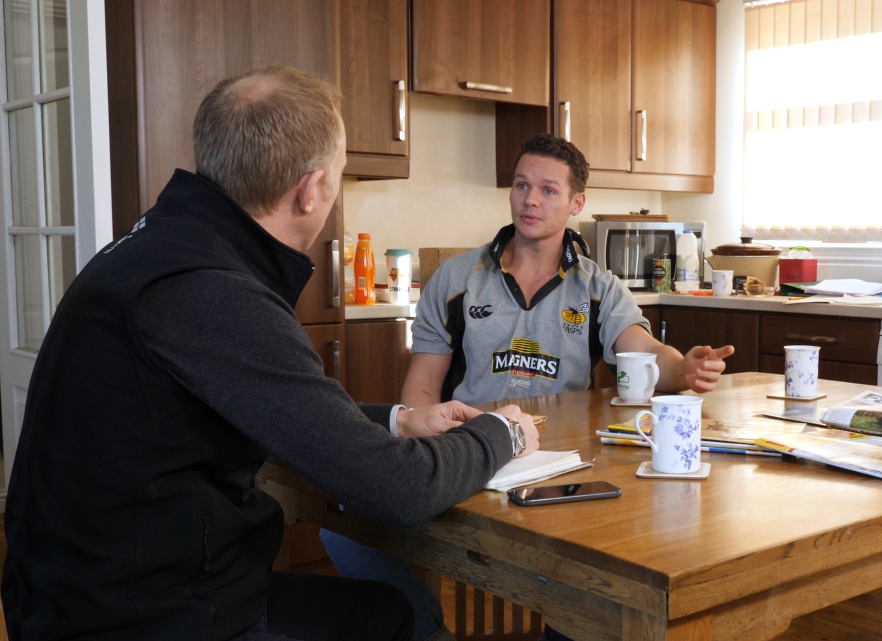 Two men gathered around a kitchen table with cups of tea, in discussion.