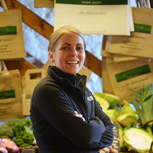 Claire Pollock standing in front of various vegetables and produce with her arms crossed, smiling