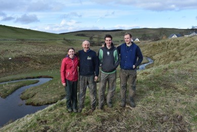 A photo showing Phillip Anderson and 3 members of his family (two sons and a daughter) who are young tree saplings whilst standing on a hilside with a meandering river behind them. The Anderson family have successfully planted native woodland on their organic hill-farm in the Scottish Borders. The trees were planted to deliver riparian benefits to the watercourses on their farm, which flow into the Oxnam water, and were part-funded through the sale of carbon as PIUs.
