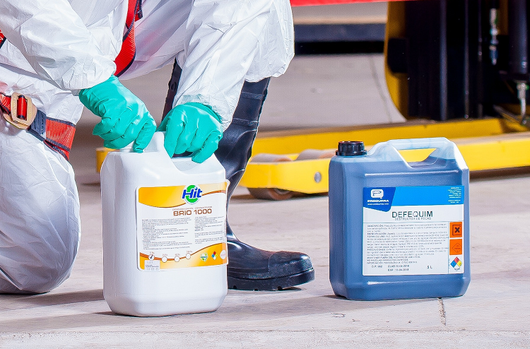 two chemical jars sitting on the floor with comprehensive labels - there is someone kneeling next to one in a white ppe suit and green nitrile gloves