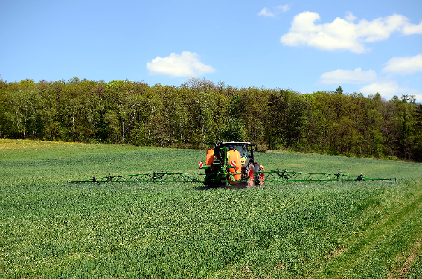 Green crop field with trees along the far boundary. Within the field is a tractor and rear mounted pesticide sprayer at work, treating the crop.