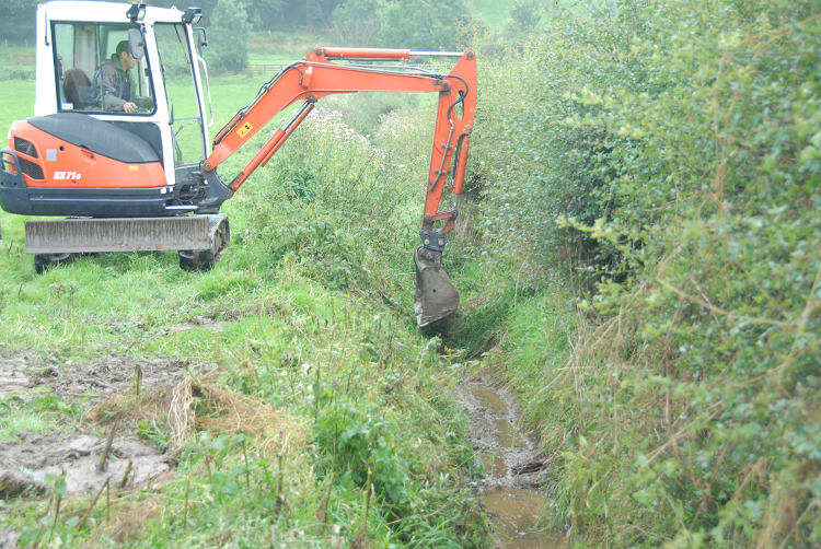 A small orange digger at work in a grassland field to clear vegetation and sediment from a boundary ditch which is adjacent to a well established hedge.