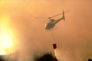 A helicopter with a water dispensing bucket below it, flying into a forest fire.