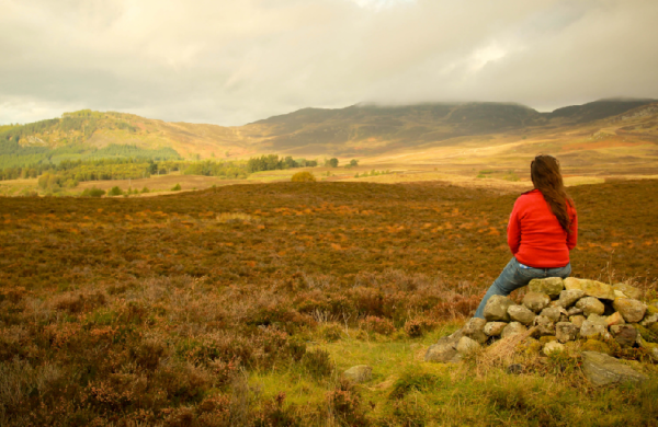 A girl sitting on a stone stack overlooking an area of peatland in the Scottish hills.