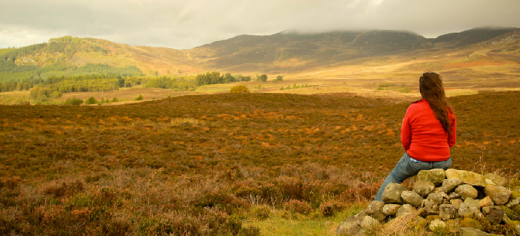 A girl sitting on a stone stack overlooking an area of peatland in the Scottish hills.