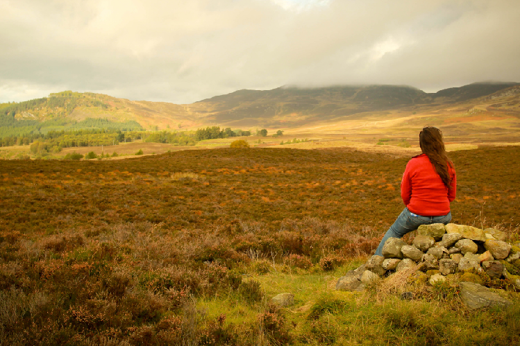 A girl sitting on a stone stack overlooking an area of peatland in the Scottish hills.