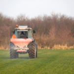 A tractor and fertiliser spreader in a grass field driving away from the camera and towards an area of scrub at the field margin.