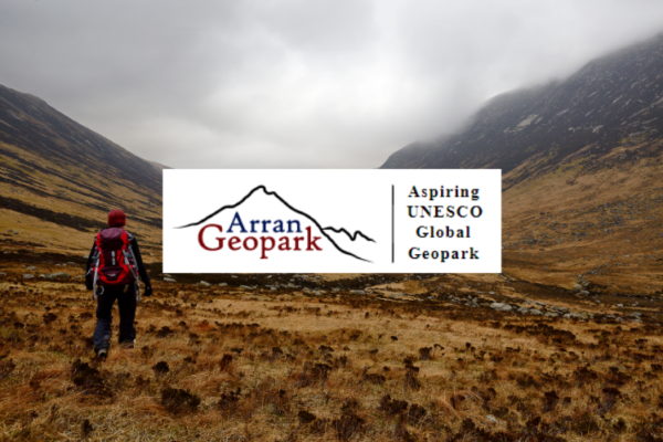 A hiker walking at the bottom of a valley on the Isle of Arran. The photo has been overlaid with the Arran Geopark logo which depicts a pencil outline of a mountain.